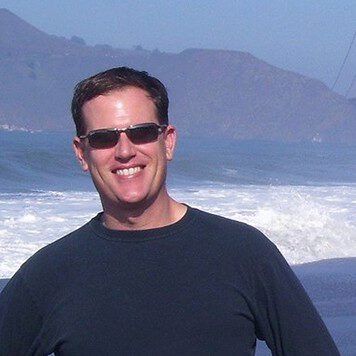 Man with dark brown hair and sunglasses standing in front of the ocean